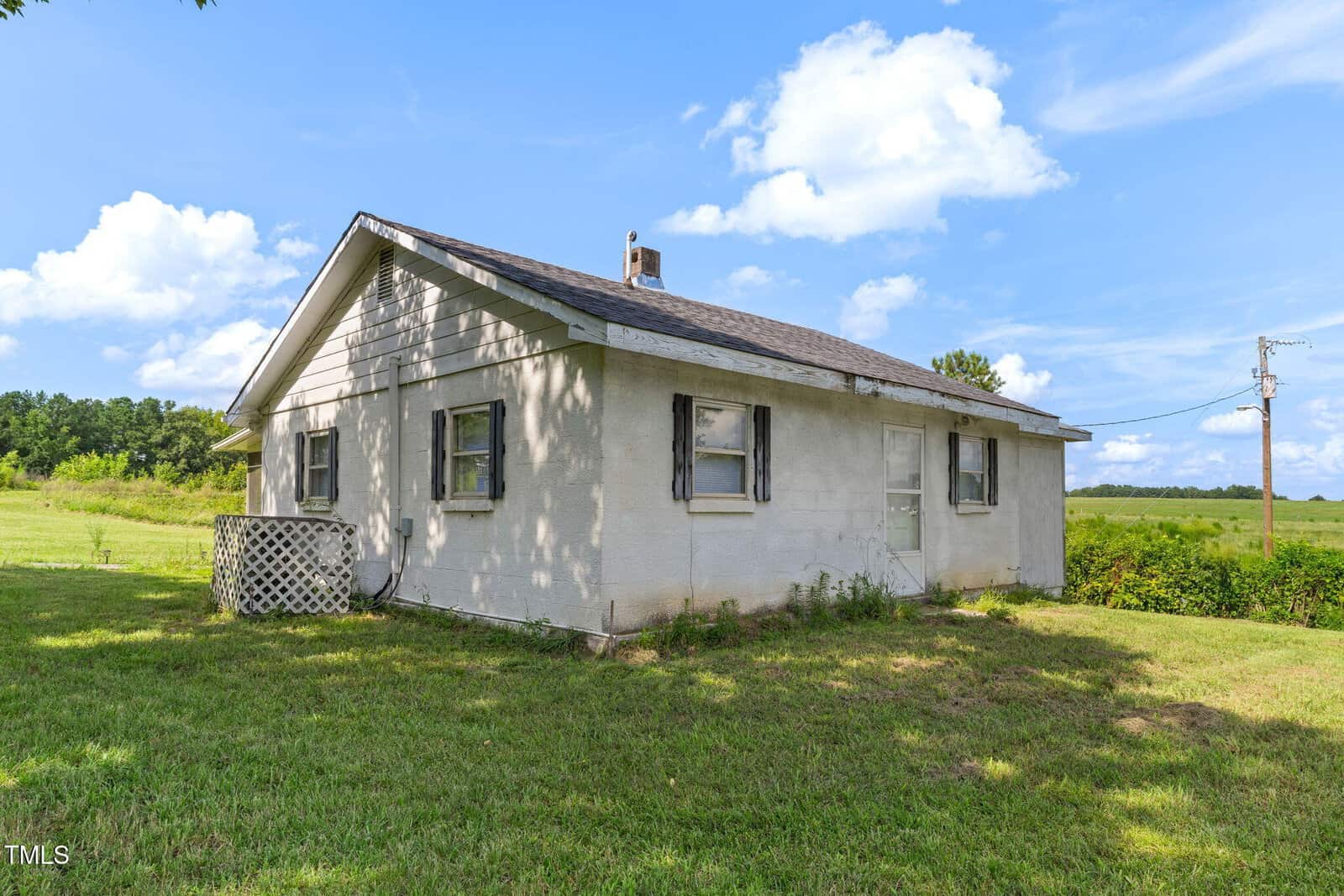 A small, single-story white house with a shingled roof and old wooden shutters, set on a grassy lot with open fields and blue sky in the background. An air conditioning unit is next to the house.