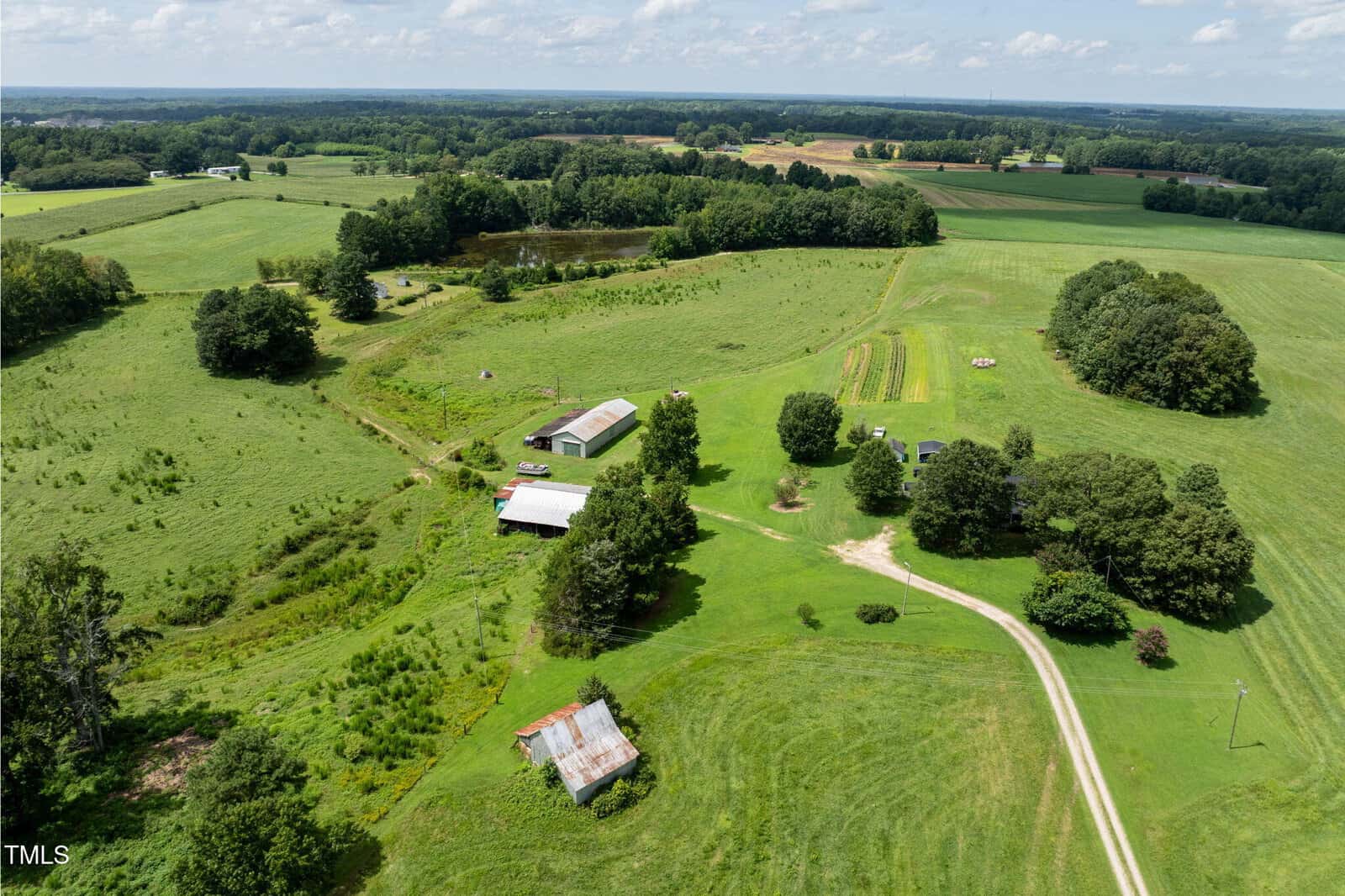 Aerial view of a rural landscape with open green fields, scattered trees, a winding dirt road, several barns and sheds, a pond, and a few small cultivated plots under a partly cloudy sky.