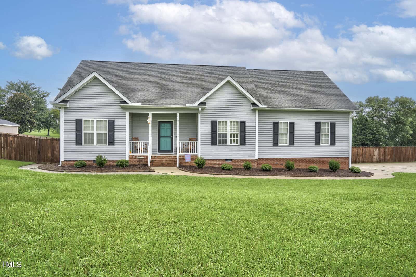 Single-story gray house with white trim, dark shutters, and a teal front door. The home sits on a large, well-maintained lawn with minimal landscaping and a wooden fence in the background under a partly cloudy sky.