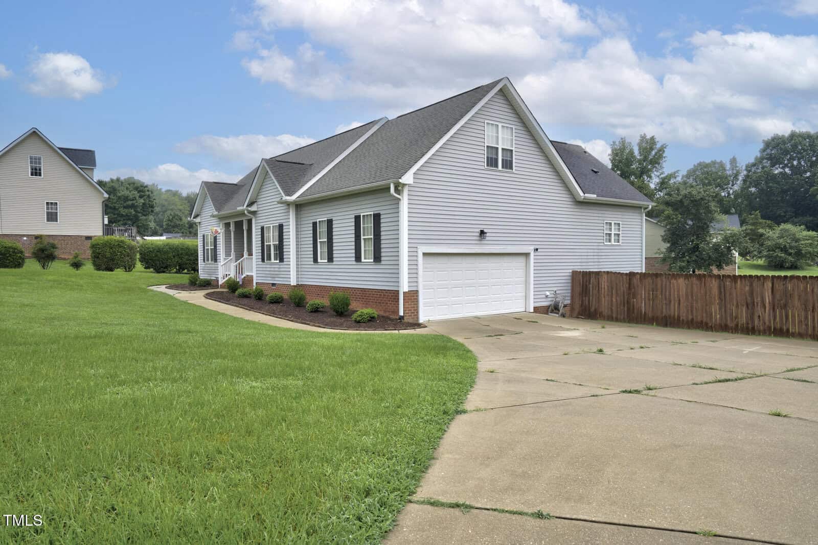 Single-story white house with dark shutters and a gray roof, attached two-car garage, brick foundation, long curved driveway, manicured lawn, and wooden fence, set on a spacious lot under a partly cloudy sky.