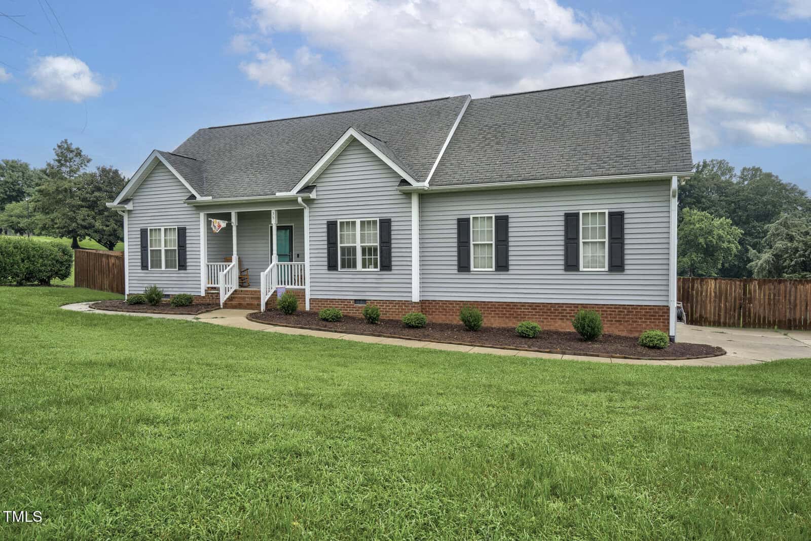 A single-story house with light gray siding, black shutters, a teal front door, and a small porch sits on a large, well-manicured lawn with shrubs and a wooden fence in the background under a partly cloudy sky.