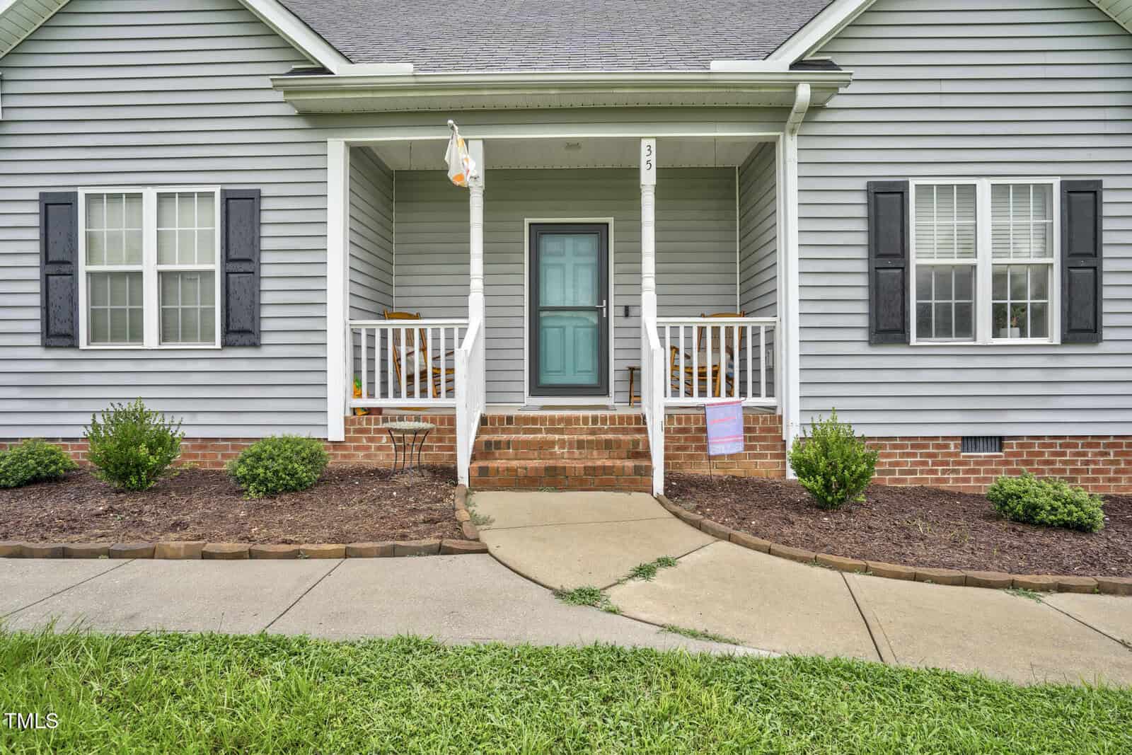 Front view of a gray house with white trim, a covered porch, two rocking chairs, a blue door, brick steps, and black shutters on the windows. Mulched flower beds with green shrubs line the walkway.