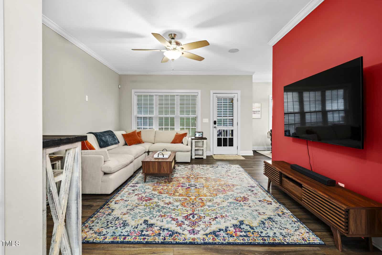 A modern living room with a beige sectional sofa, colorful pillows, a patterned rug, a wall-mounted TV on a red accent wall, wood floors, and large windows with white blinds. A ceiling fan hangs from the white ceiling.