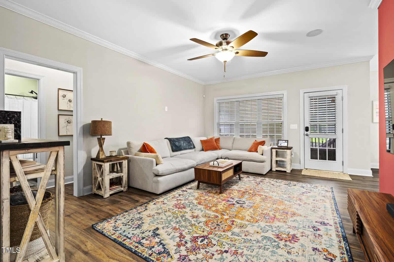 A living room with a beige sectional sofa, colorful patterned rug, wooden coffee table, and side tables. A ceiling fan hangs overhead. There is a door with blinds, large windows, and wood flooring. Walls are light with red accent visible.