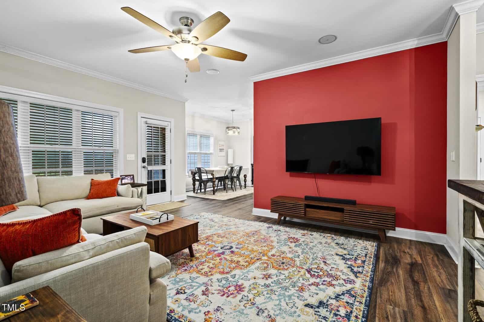 A living room with a beige couch, chair, and colorful rug faces a wall-mounted TV on a red accent wall. A ceiling fan hangs above, and a dining area is visible in the background.