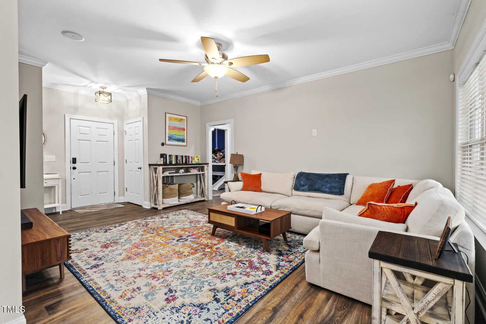 Bright living room with a beige sectional sofa, colorful pillows, a patterned area rug, wooden coffee table, TV stand, and console table near a white front door. Ceiling fan and windows provide light. Artwork hangs on the wall.