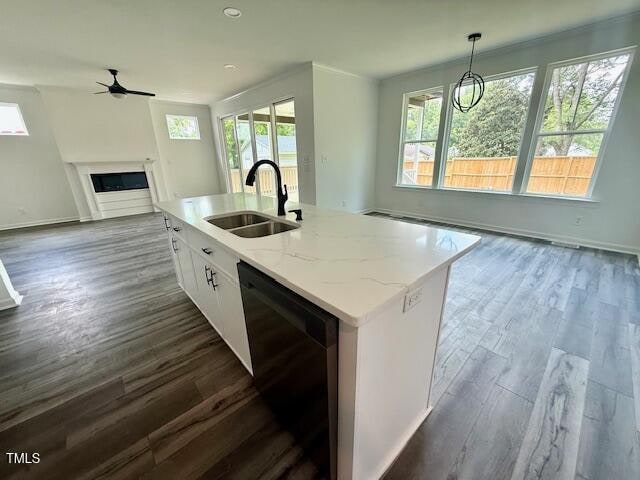 A modern open-concept kitchen and living area with a white island, stainless steel sink, dishwasher, wood-look flooring, large windows, and a fireplace in the background. A dining area features a black pendant light.