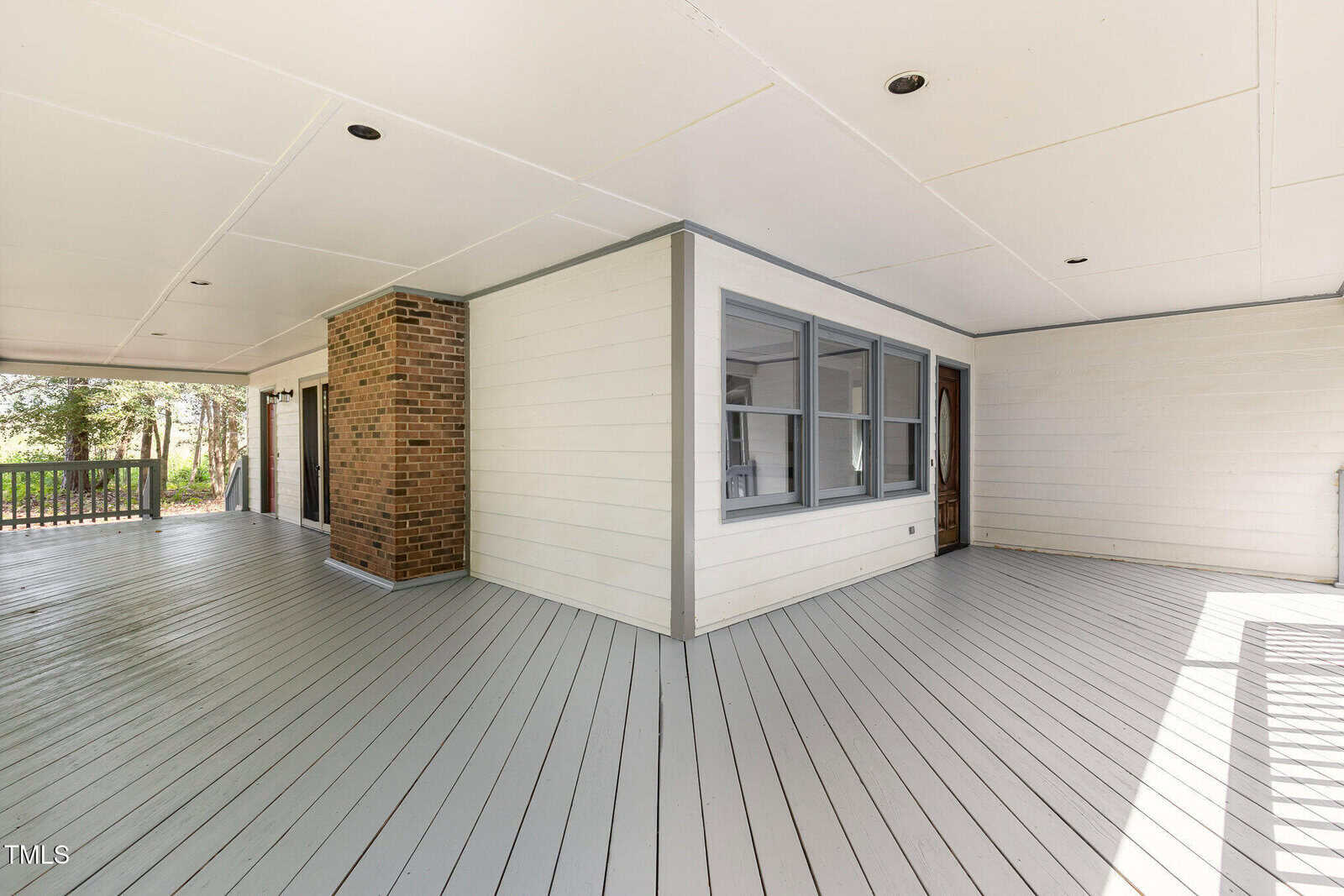 A spacious covered porch with light gray wooden flooring, white siding walls, a brick column, large windows, and recessed ceiling lights. A railing and greenery are visible in the background.
