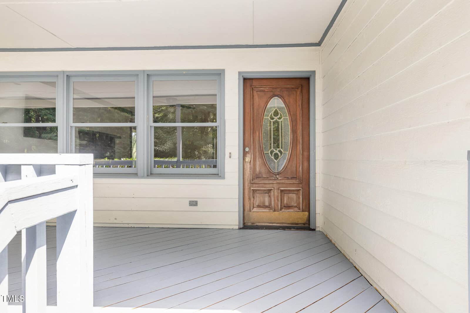 A covered porch with light gray flooring and cream-colored siding, featuring a wooden front door with an oval glass insert and three large windows to the left of the door.