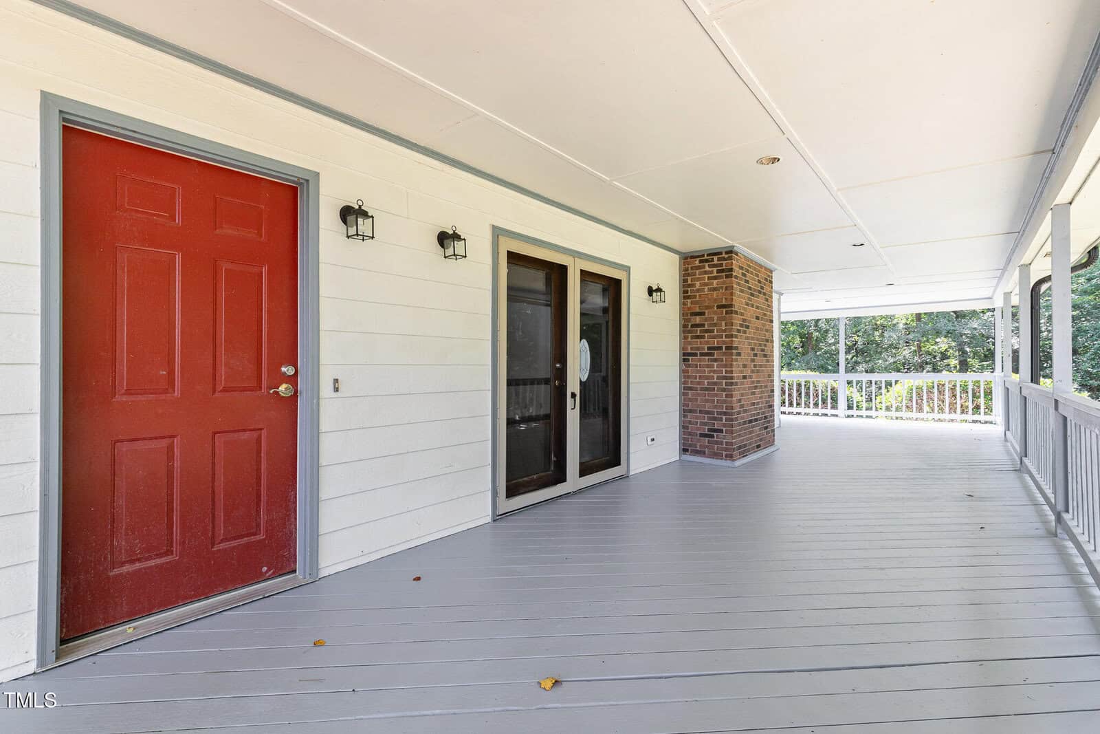 A spacious covered porch with gray flooring, white siding, a red door, two black lantern lights, double glass doors, a brick column, and a white railing overlooking a green, wooded area.