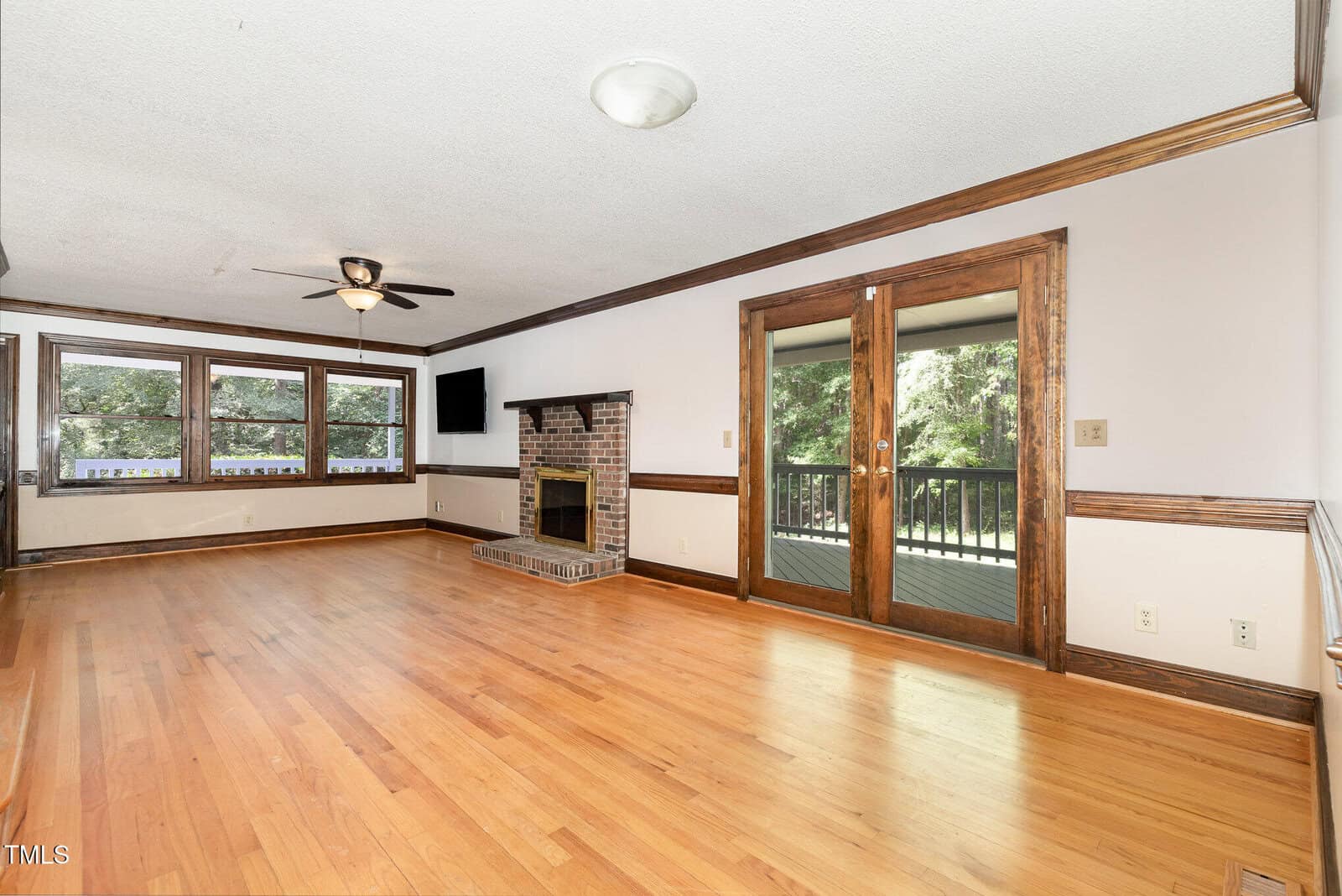 Spacious living room with hardwood floors, a brick fireplace, ceiling fan, large windows, and double glass doors leading to a covered porch with views of trees outside.