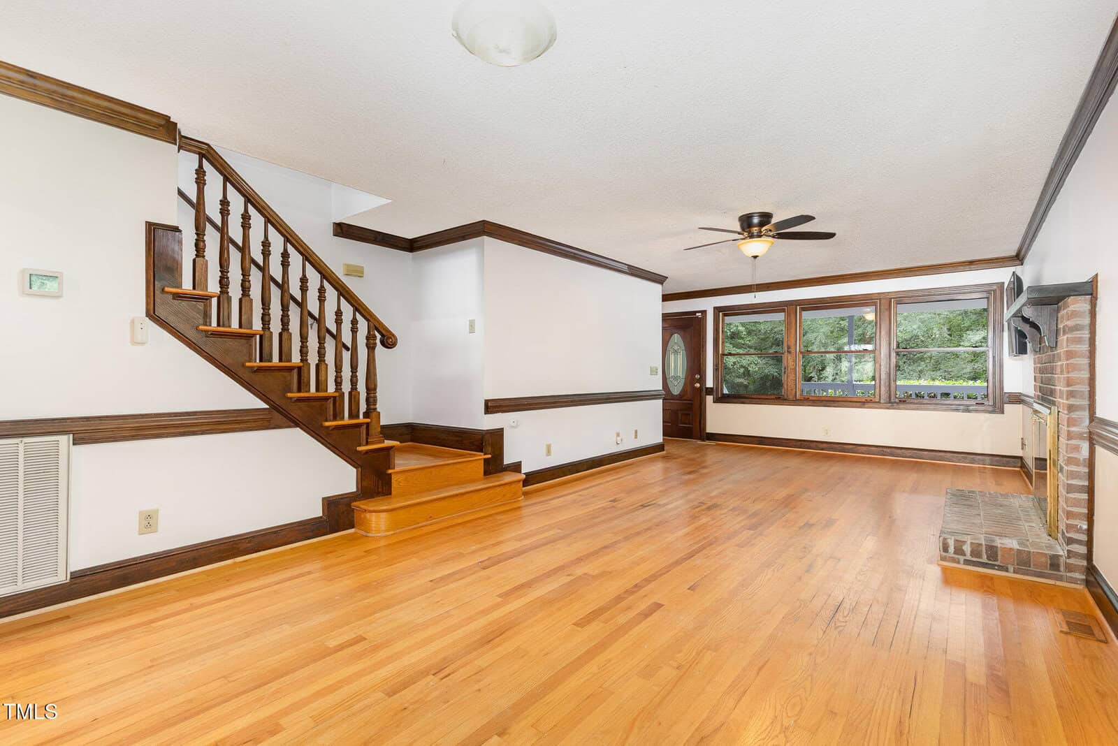 Spacious living room with hardwood floors, wood trim, ceiling fan, brick fireplace, large windows, and a wooden staircase leading to the upper floor. Walls and ceiling are painted white.