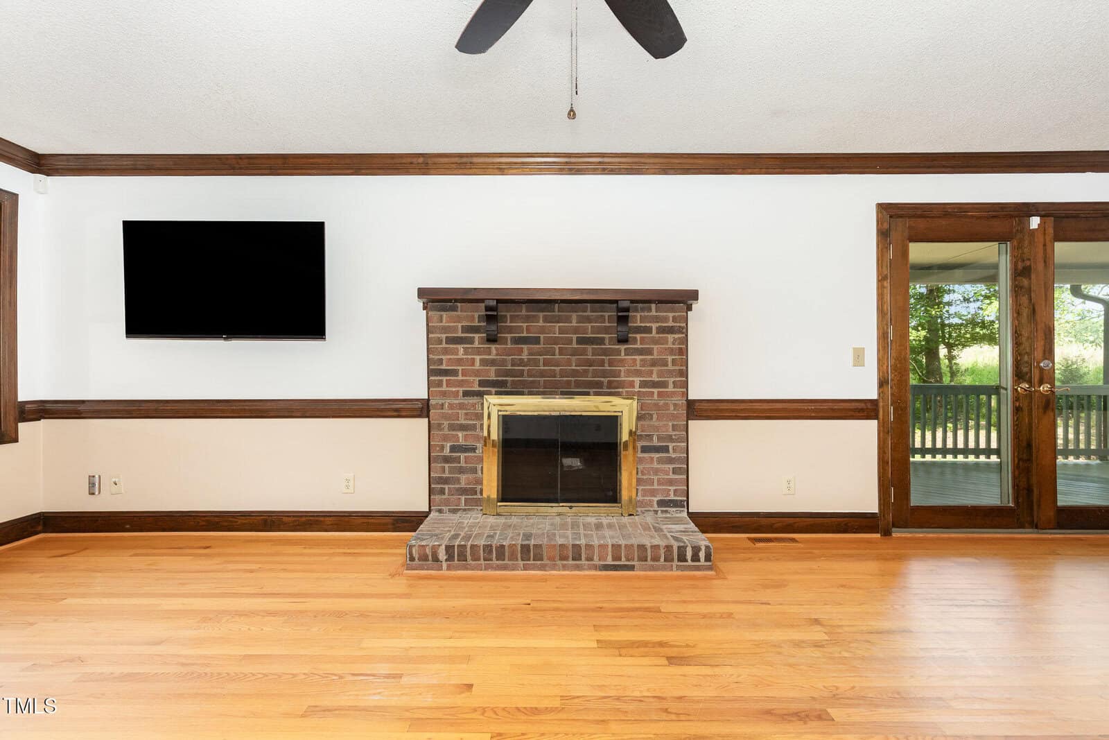 A brick fireplace with a brass-trimmed glass door is centered on a white wall. A flat-screen TV is mounted to the left. Hardwood floors and wood trim are visible, along with double glass doors to a deck on the right.