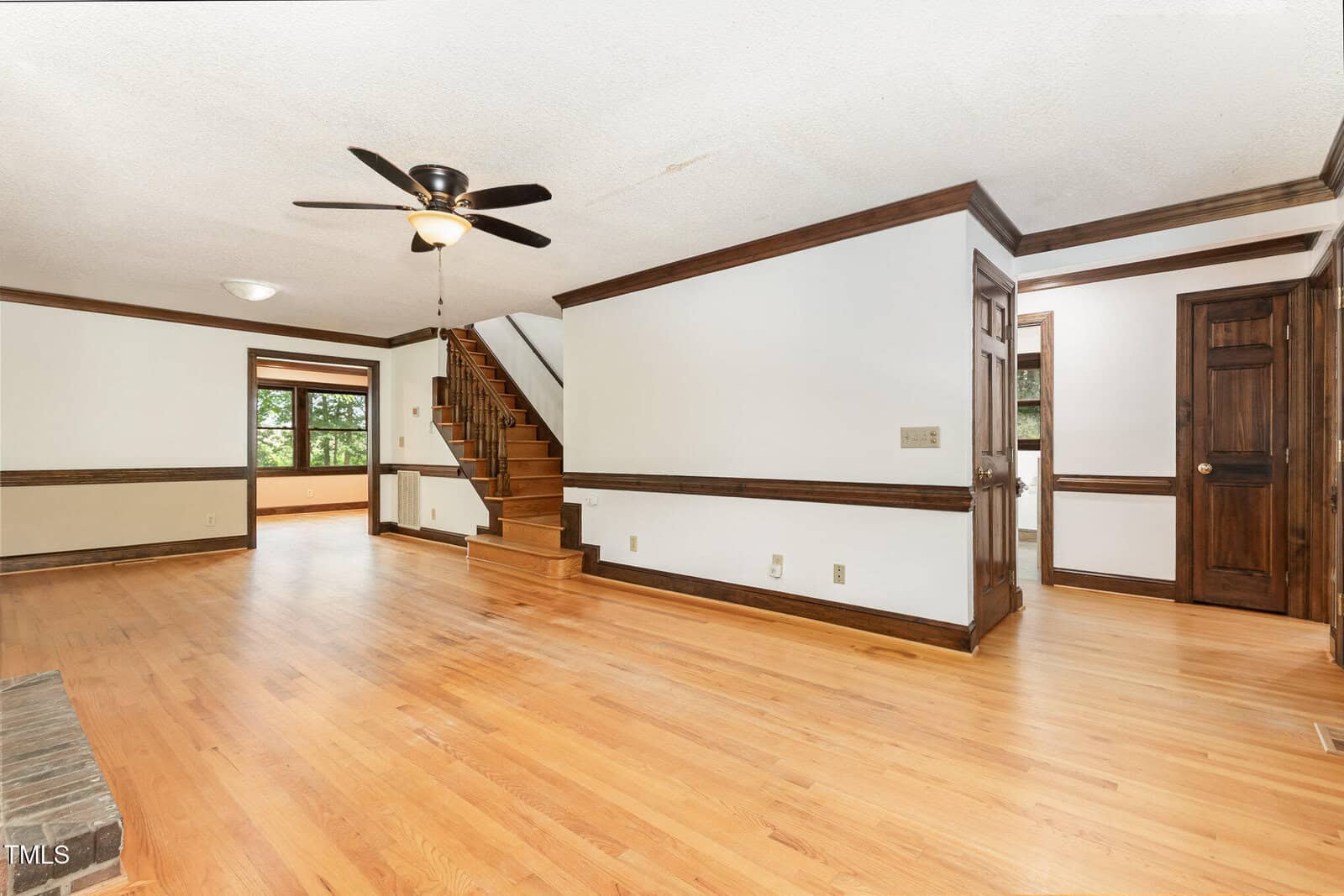 Spacious living room with hardwood floors, white walls, dark wood trim, a ceiling fan, a staircase, and multiple doors. Large window lets in natural light. Room connects to additional spaces in the home.