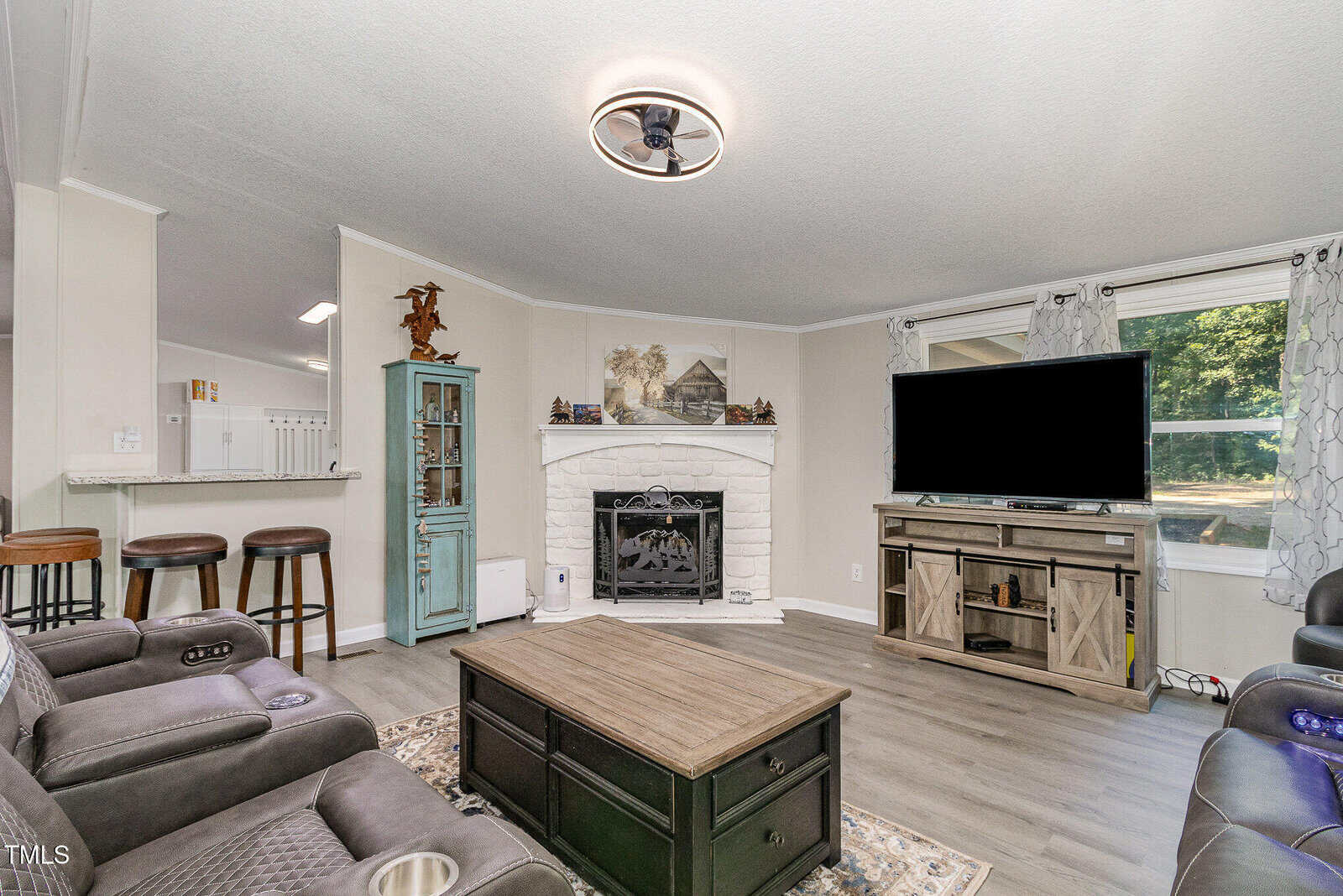 A living room with gray recliners, a wooden coffee table, and a TV on a wooden stand. There is a white brick fireplace with a mantel and artwork above it, a green cabinet, and windows with sheer curtains letting in daylight.