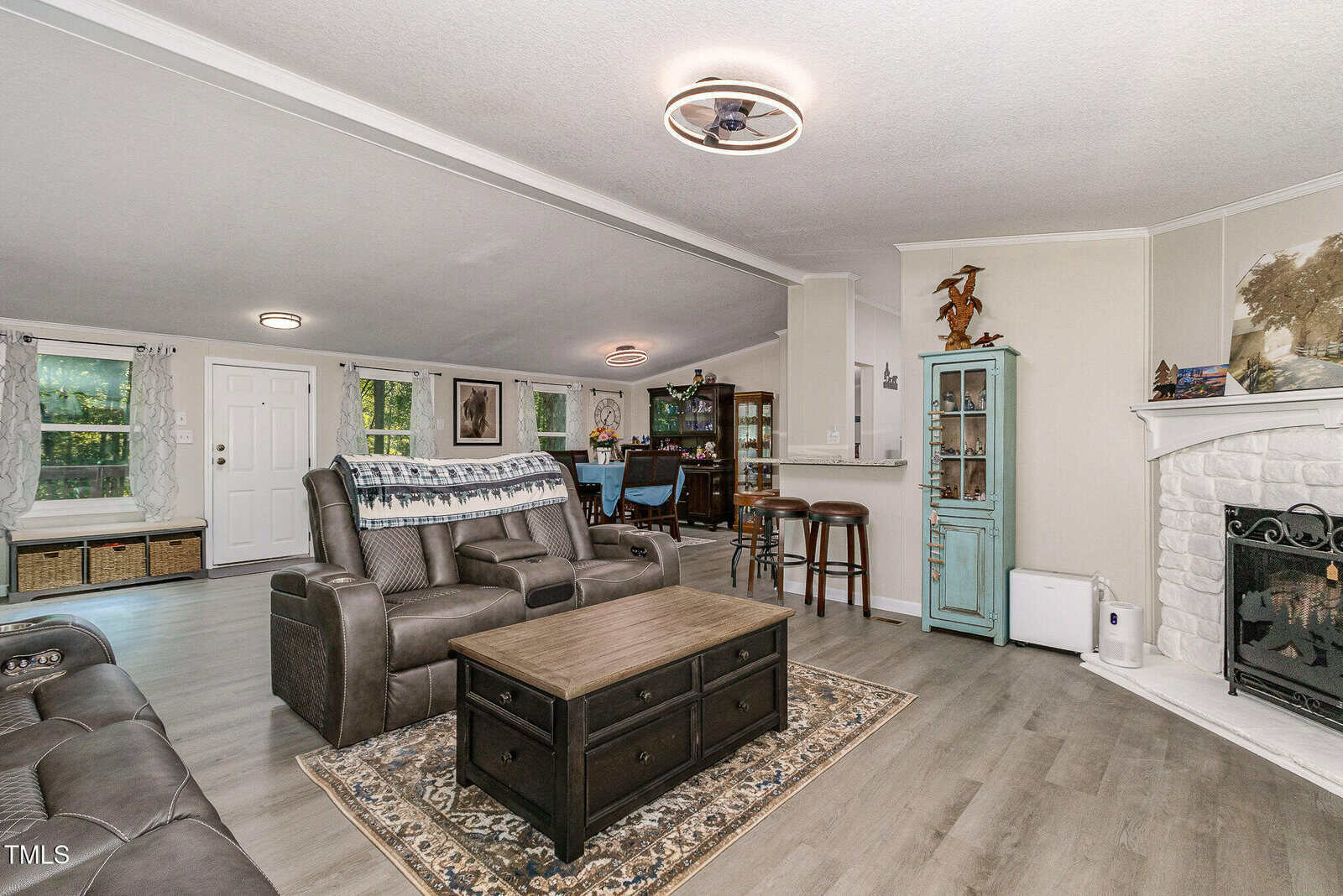 A spacious living room with gray wood flooring, brown leather seating, a coffee table on a rug, a fireplace on the right, a blue cabinet, and a dining area with a wooden table and chairs in the background.
