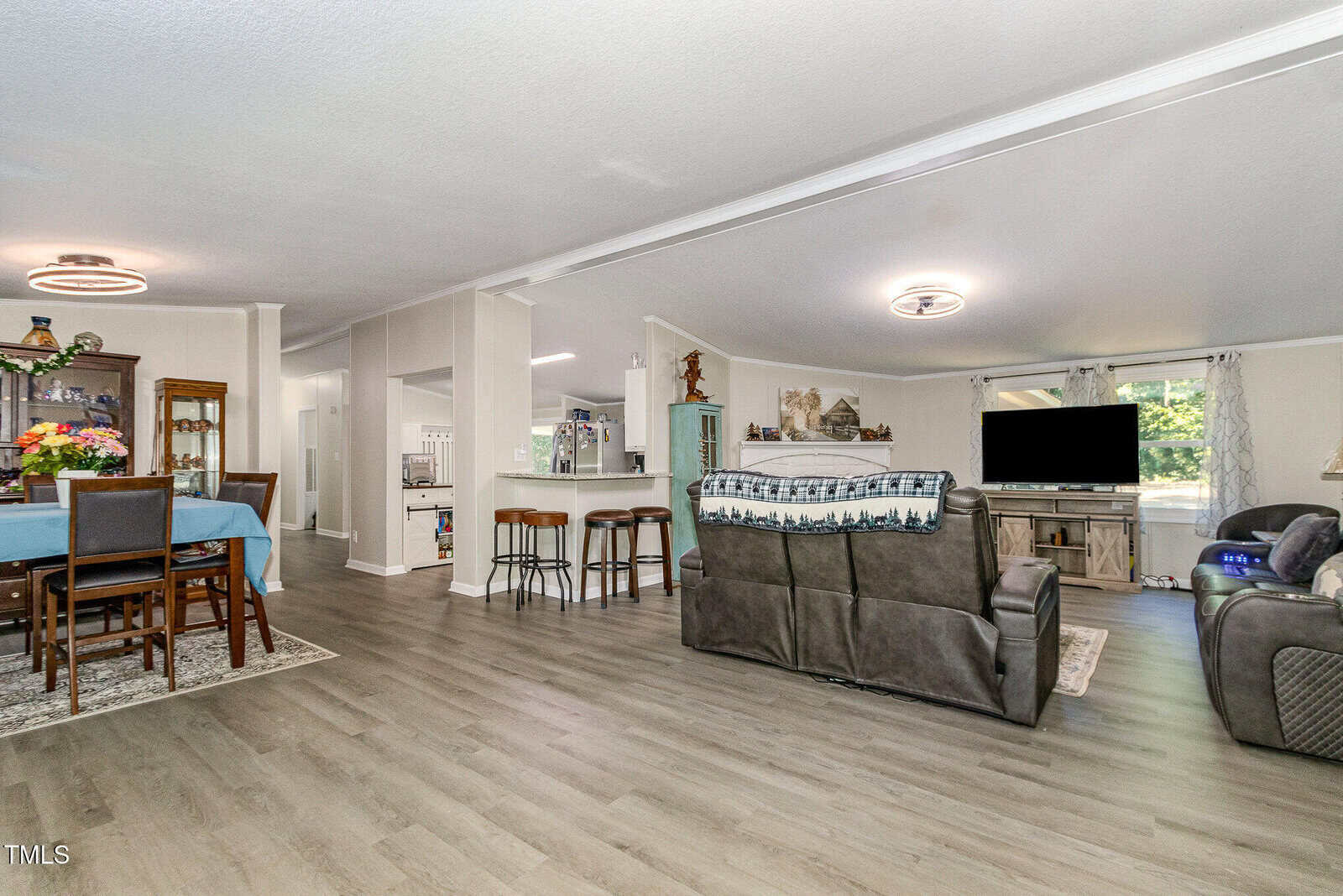 Open-concept living area with gray wood flooring, a dining table with blue tablecloth, a kitchen with bar seating, and a living room featuring sofas, a large TV, and a window letting in natural light.