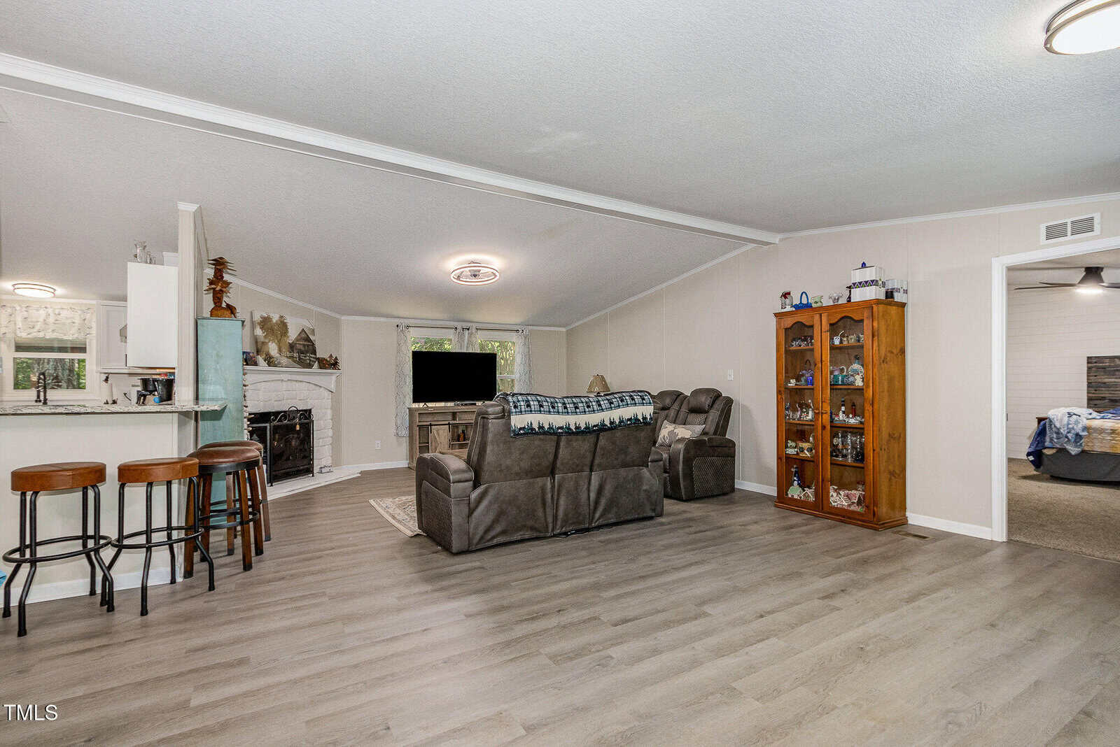 A living room with gray flooring, a brown reclining sofa, two display cabinets, and a TV; an open kitchen with bar stools is visible on the left, and a bedroom is partially seen through a doorway on the right.