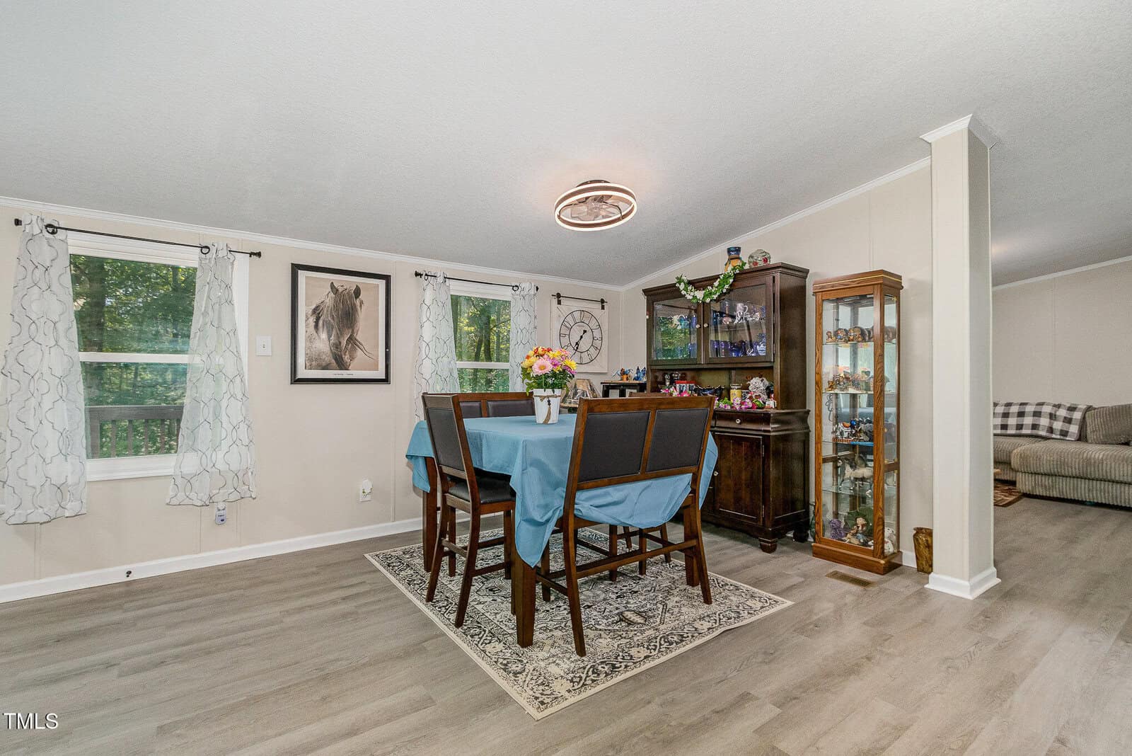 A dining area with a table covered by a blue cloth, six chairs, a floral centerpiece, a glass-front cabinet, wall art, and large windows with patterned curtains; adjacent to a living space.