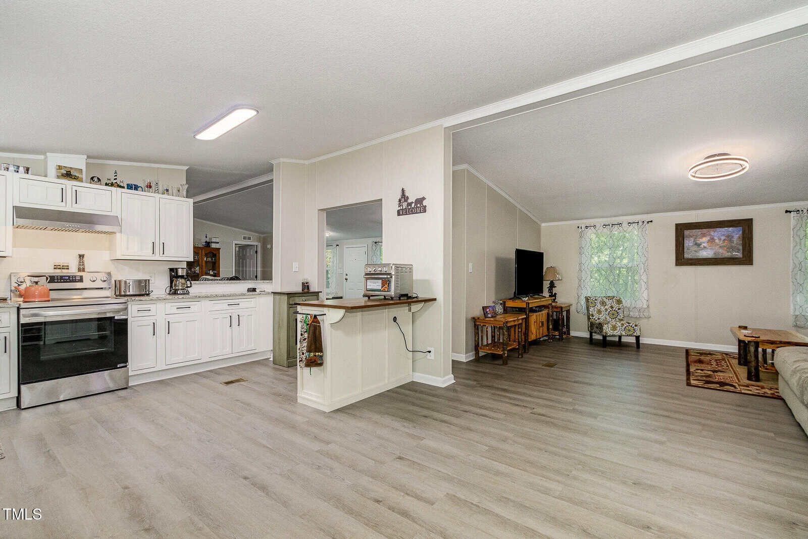 Open-plan living area with light wood flooring, white kitchen cabinets, a stainless steel stove, a breakfast bar, and a living room featuring a sofa, armchair, TV, and natural light from large windows.
