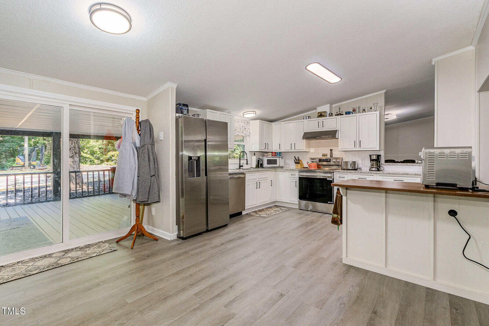 A modern kitchen with white cabinets, stainless steel appliances, and light wood flooring. A coat rack with two aprons is next to sliding glass doors that lead to a screened porch with outdoor seating.