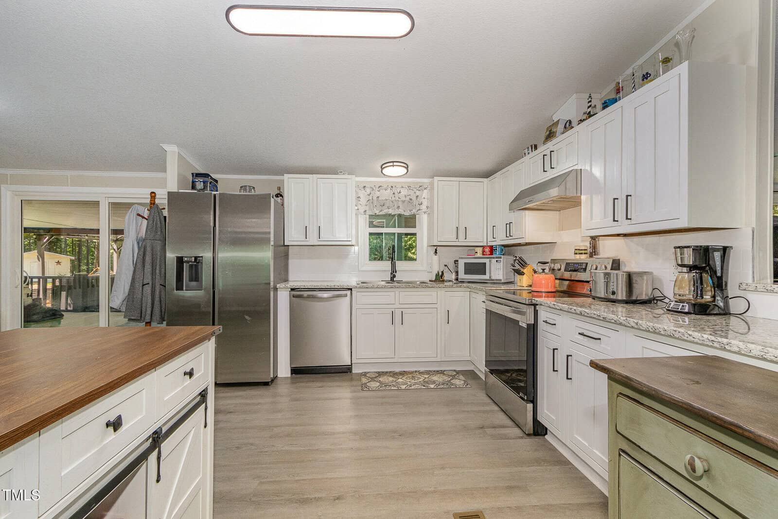 A modern kitchen with white cabinets, stainless steel appliances, a double-door refrigerator, oven, dishwasher, microwave, coffee maker, and a window above the sink. An island is in the foreground and hardwood floors throughout.