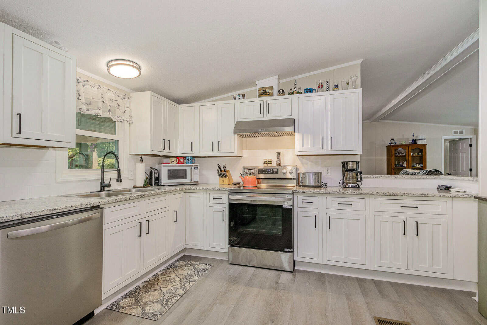 Modern kitchen with white cabinets, stainless steel appliances, a patterned rug in front of the oven, a coffee maker, microwave, and decorative items on top of the cabinets. A window above the sink lets in natural light.