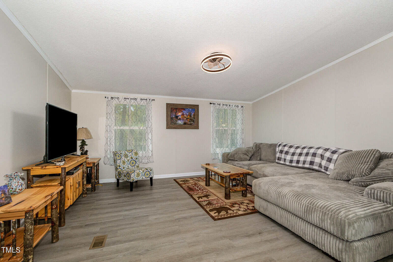 A cozy living room with a gray sectional sofa, patterned chair, wooden tables, TV on a stand, decorative rug, and curtains on windows. A painting hangs on the back wall above the sofa.