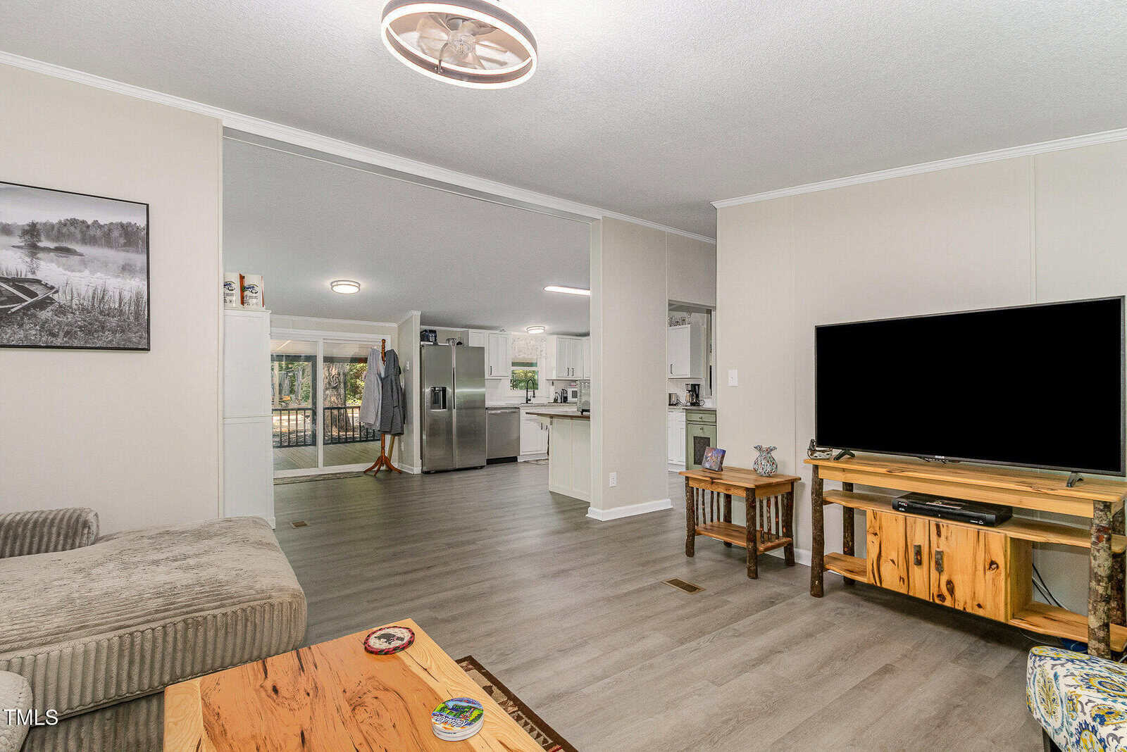 Modern living room with a TV on a wooden stand, a striped sectional sofa, a wooden coffee table, and a view into a bright kitchen with white cabinets and stainless steel appliances.