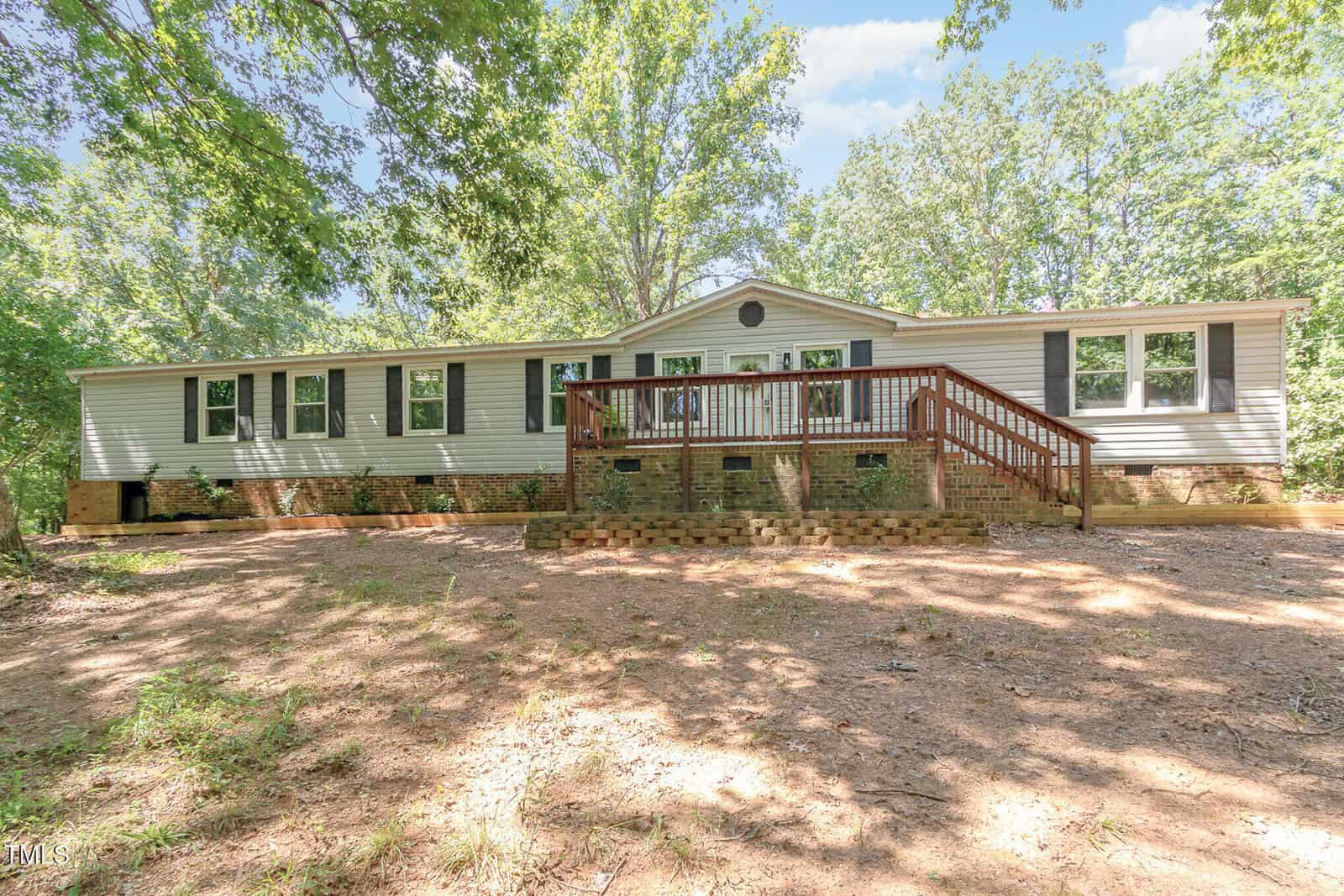 A single-story beige manufactured home with black shutters and a large wooden front deck at 4032 Royal Oak Drive, Franklinton, surrounded by trees and dappled sunlight on a dirt yard.