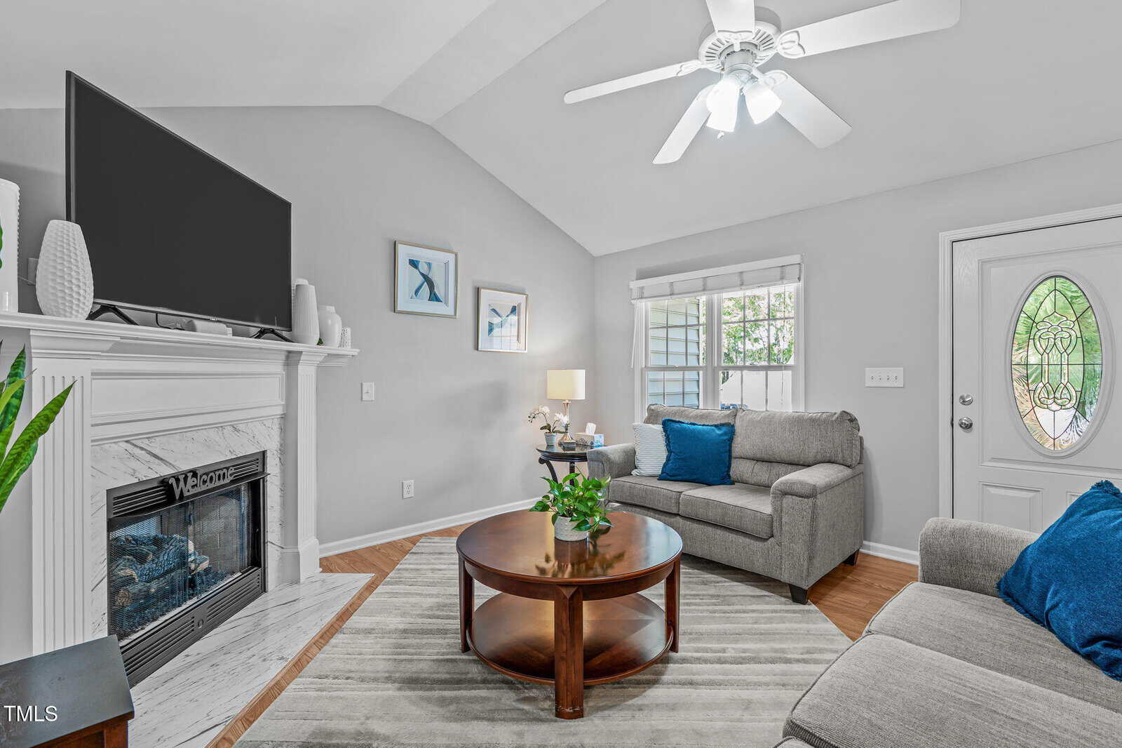 A living room with light gray walls, a ceiling fan, a fireplace with a TV above it, two gray armchairs with blue pillows, a round wooden coffee table, and a door with an oval window.