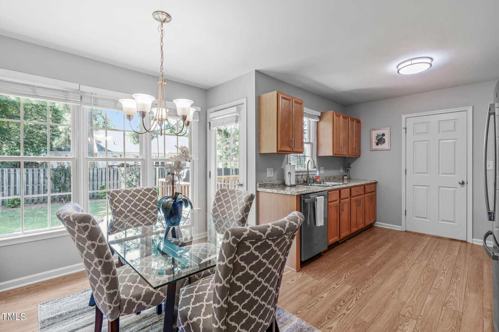A kitchen and dining area with light wooden floors, a glass dining table with four patterned chairs, wooden cabinets, stainless steel appliances, large windows, and a door leading outside.