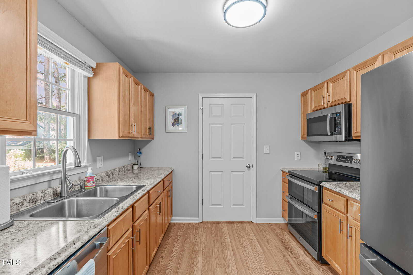 A modern kitchen with light wood cabinets, granite countertops, stainless steel appliances, a double sink under a window, and a door at the center of the back wall. The floor is a light wood laminate.