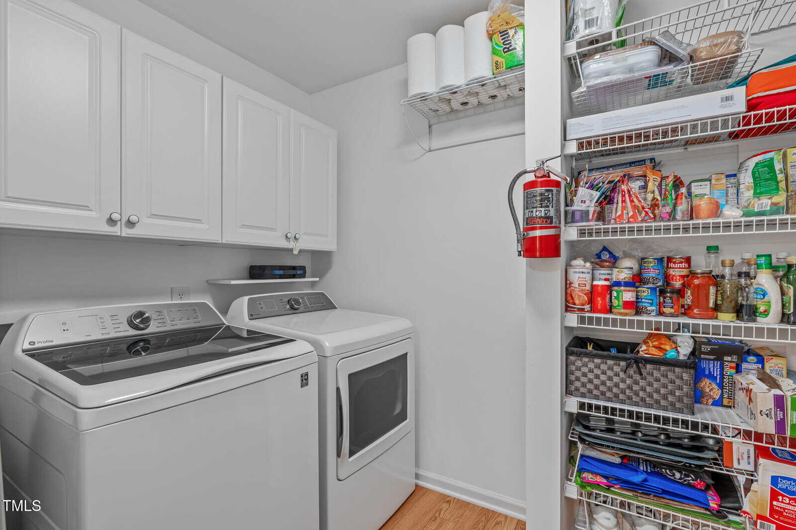 A laundry room with a washer and dryer on the left, white cabinets above, and a pantry shelf on the right holding food, paper towels, a fire extinguisher, and cleaning supplies.