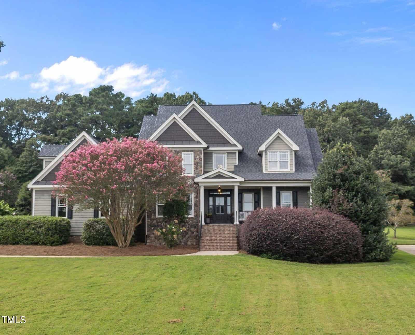 Charming two-story house at 240 Barnhill Lane, Wake Forest, featuring gray siding, white trim, and a dark shingled roof. Flowering trees and bushes grace the landscaped yard with a well-manicured lawn, all set against a backdrop of tall green trees under a blue sky.