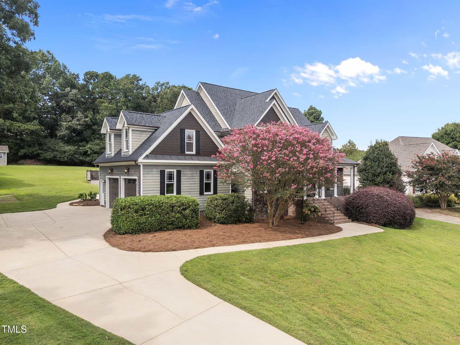 A two-story gray house with a gabled roof, attached garage, and brick stairs leading to the front door. The yard has neatly trimmed grass, a curved driveway, and flowering trees and shrubs.