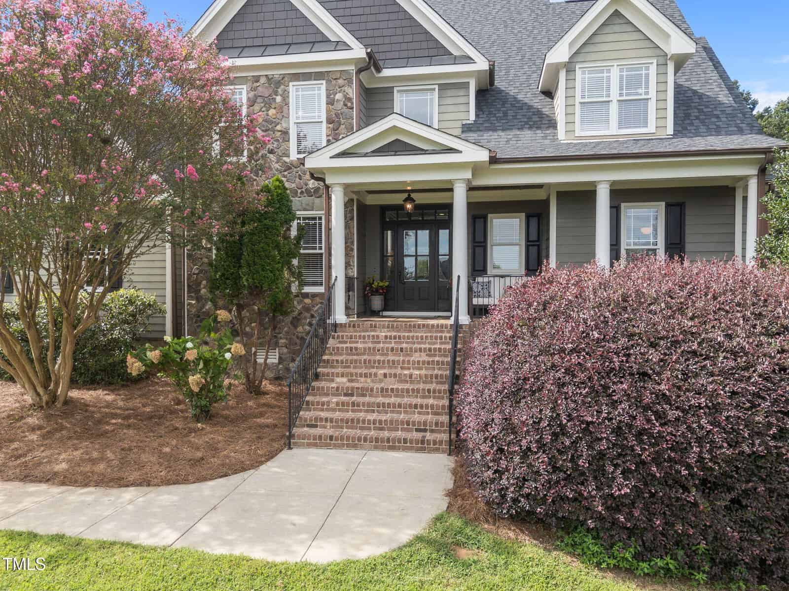 Two-story house with gray siding, brick steps leading to a covered front porch, black double doors, white trim, a stone accent wall, and landscaped bushes and trees in the front yard.