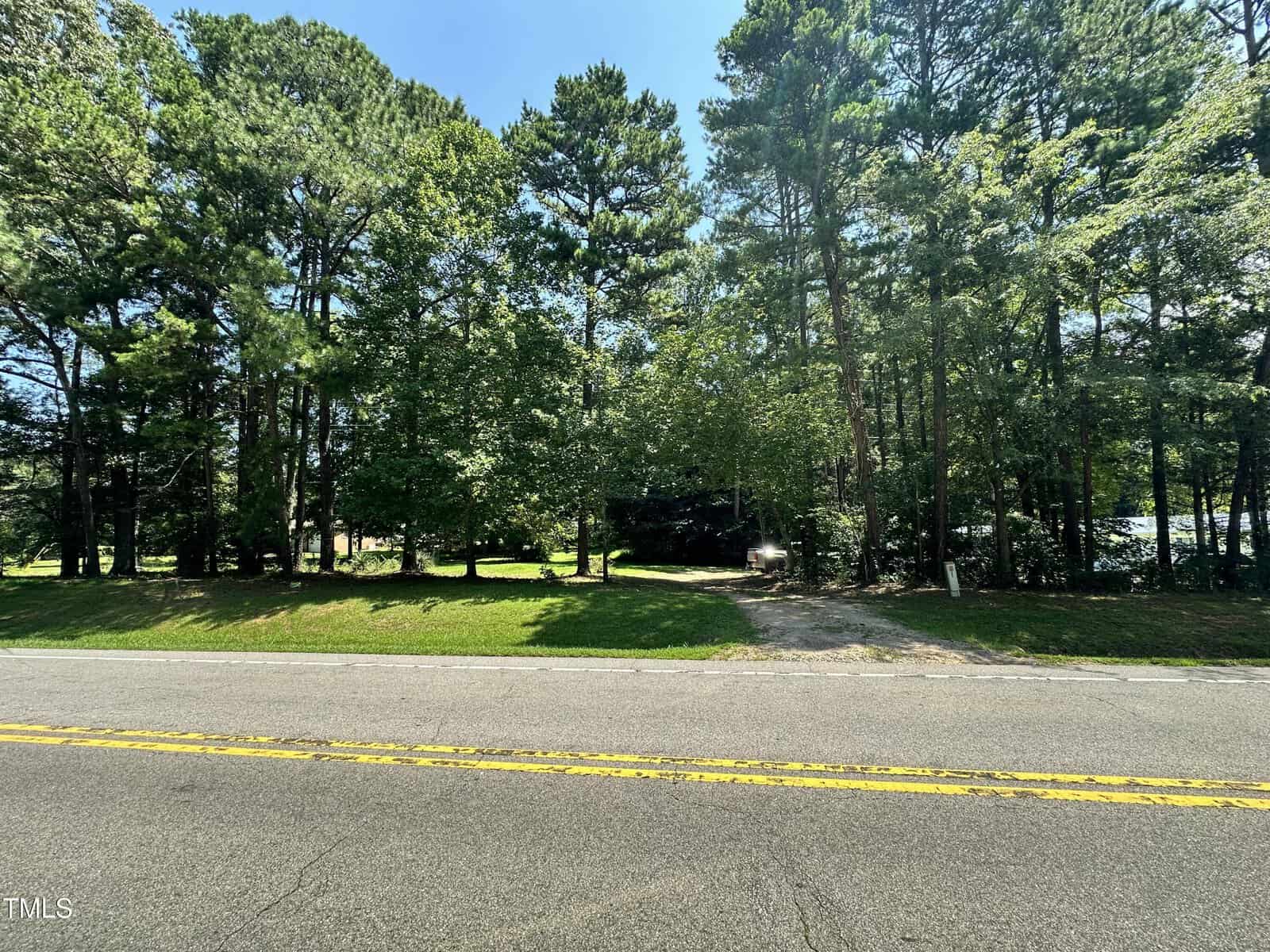 View of a paved road with double yellow lines in Henderson, bordered by grass and dense trees. A gravel driveway leads into the wooded 0 Tom Hicks Land, as sunlight filters through the branches.