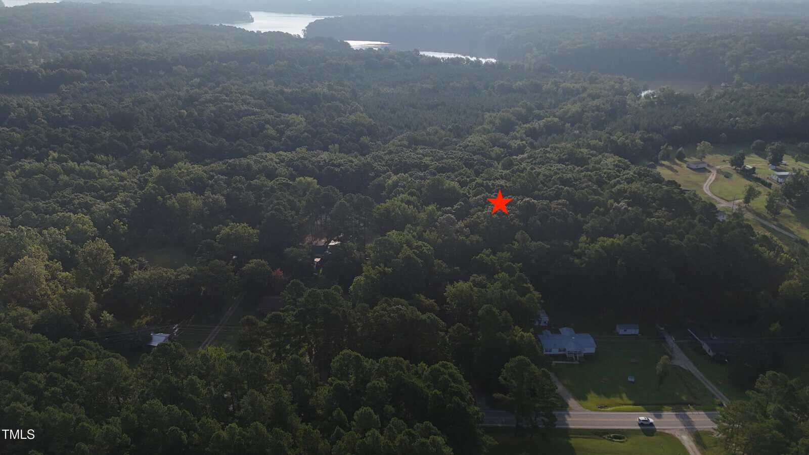 Aerial view of a wooded area with a few houses and a winding road in the foreground. A red star marks a specific spot among the trees. A large lake is visible in the background.