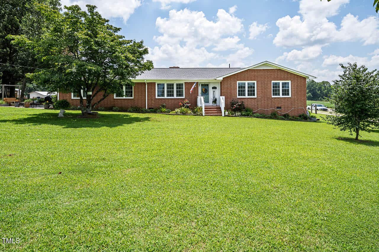Charming single-story brick house with white trim and a gray roof at 2526 Warrenton Road in Henderson. Set on a large, well-maintained grassy lawn with trees, shrubs, and a partly cloudy sky in the background.