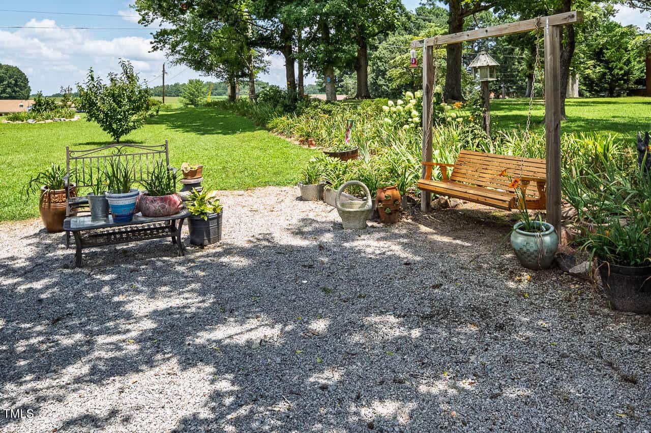 A gravel patio area with a black metal bench and cushions, several potted plants, a wooden swing on a frame, and a variety of garden decorations, surrounded by grass and trees on a sunny day.