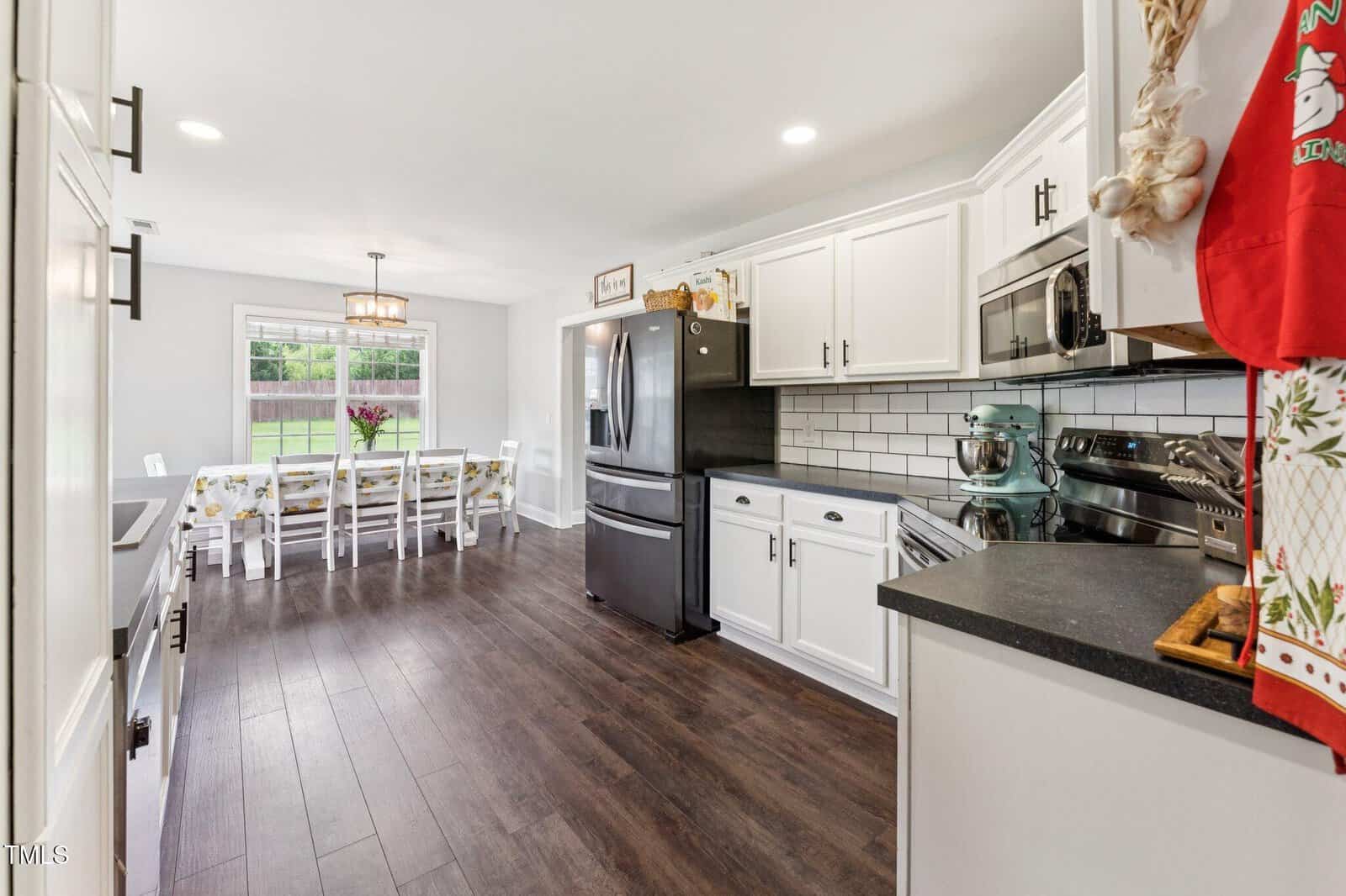 Bright kitchen with white cabinets, stainless steel appliances, dark countertops, and dark wood floors. In the background, a dining table with chairs is positioned near a large window overlooking a green lawn.