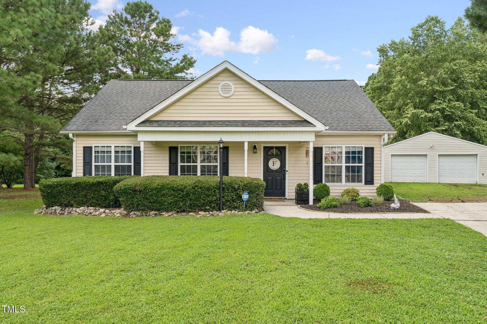 Single-story house with beige siding, dark shutters, and a gray shingled roof, surrounded by green lawn and bushes. A detached white garage is visible in the background to the right.