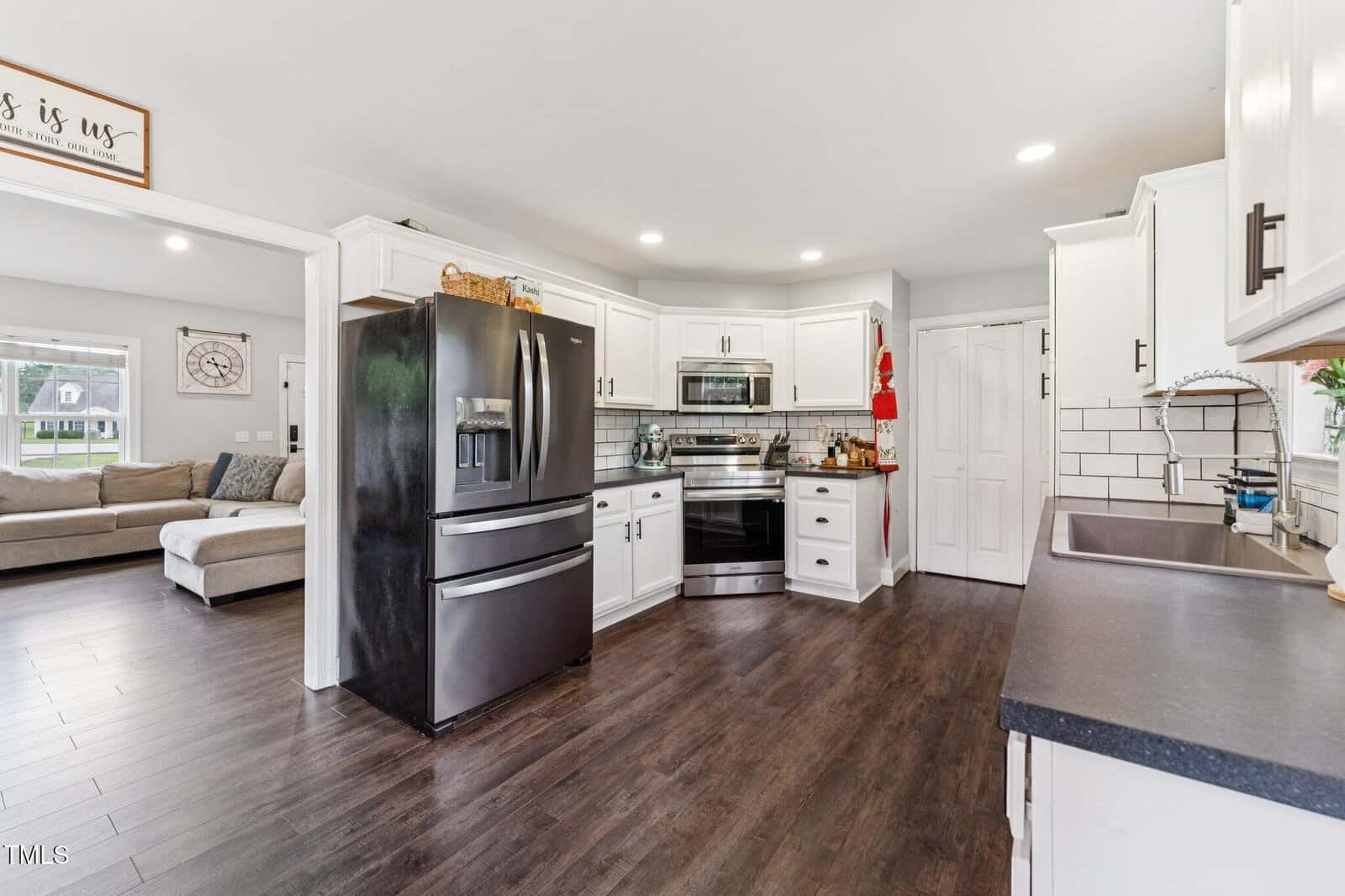 Modern kitchen with white cabinets, stainless steel appliances, dark countertops, and dark wood floors. An open doorway leads to a living room with a beige sectional sofa and large windows. Bright and clean atmosphere.
