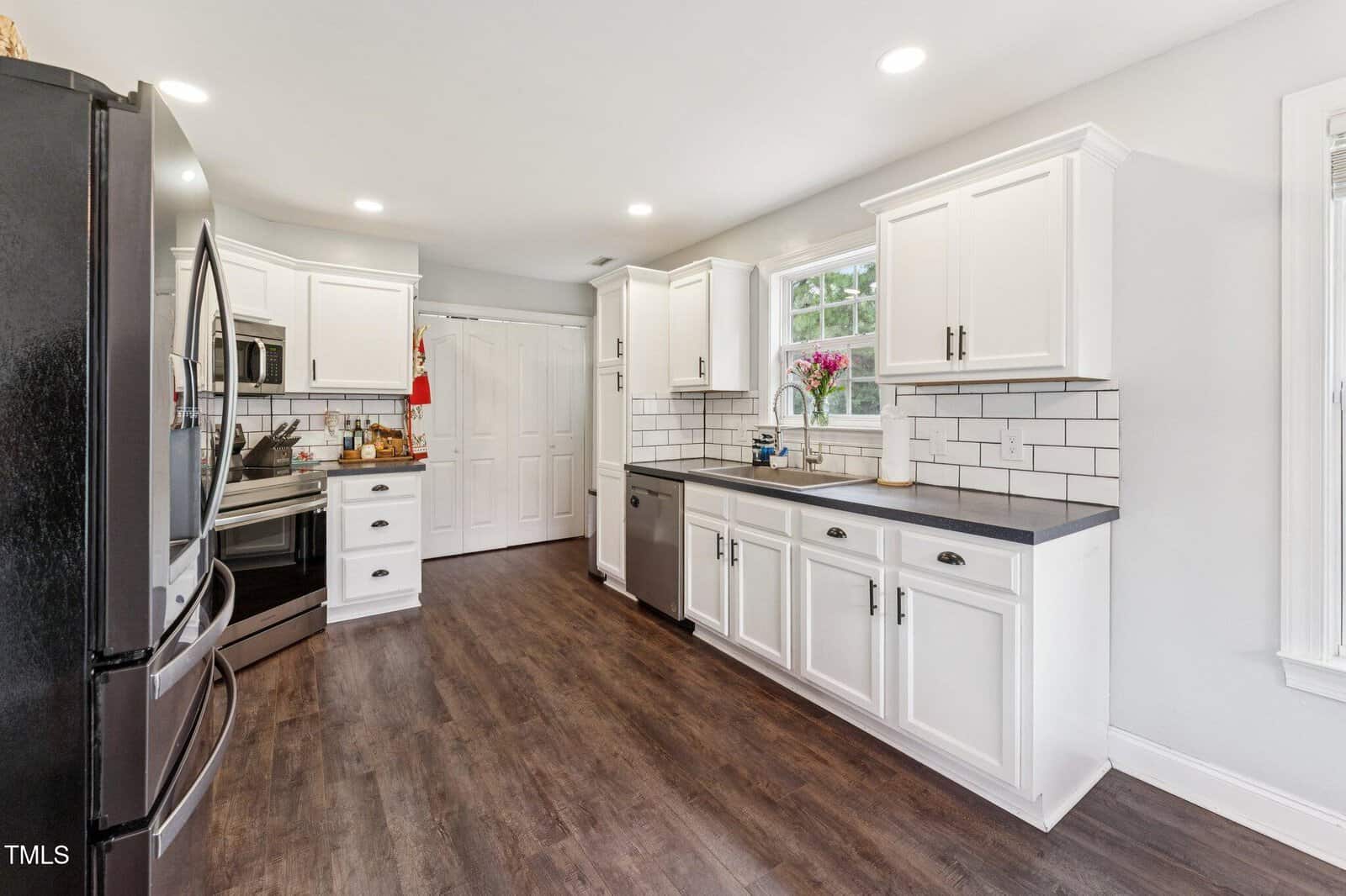 Modern kitchen with white cabinets, black countertops, stainless steel appliances, subway tile backsplash, dark wood flooring, and a window above the sink with a vase of pink flowers. Room appears bright and spacious.