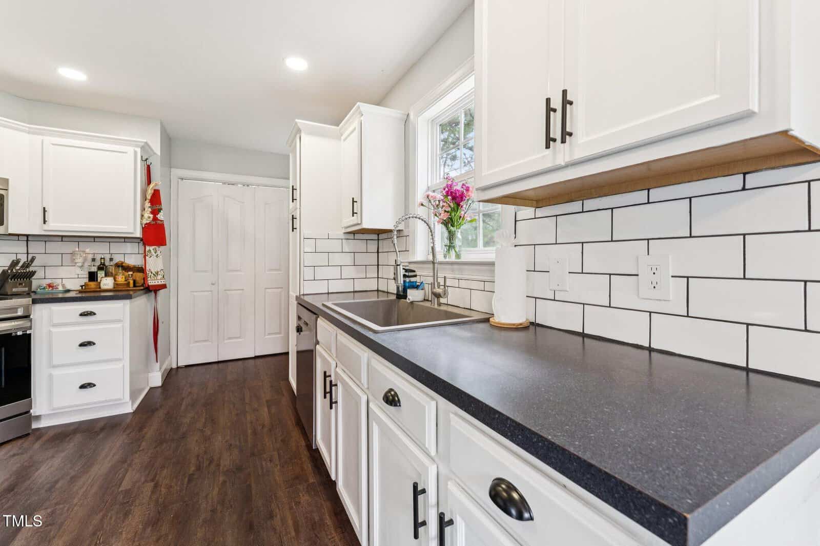 Modern kitchen with white cabinets, black countertops, subway tile backsplash, stainless steel appliances, dark wood flooring, a window above the sink, and a vase of pink flowers on the windowsill.