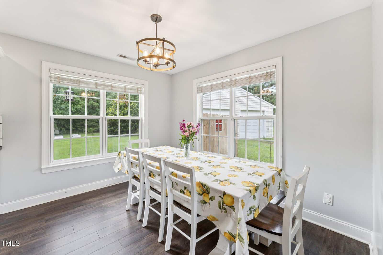 Bright dining area with two large windows, a table covered with a lemon-patterned tablecloth, six white chairs, a vase of flowers, light gray walls, and dark wood flooring. A shed and green yard are visible outside.