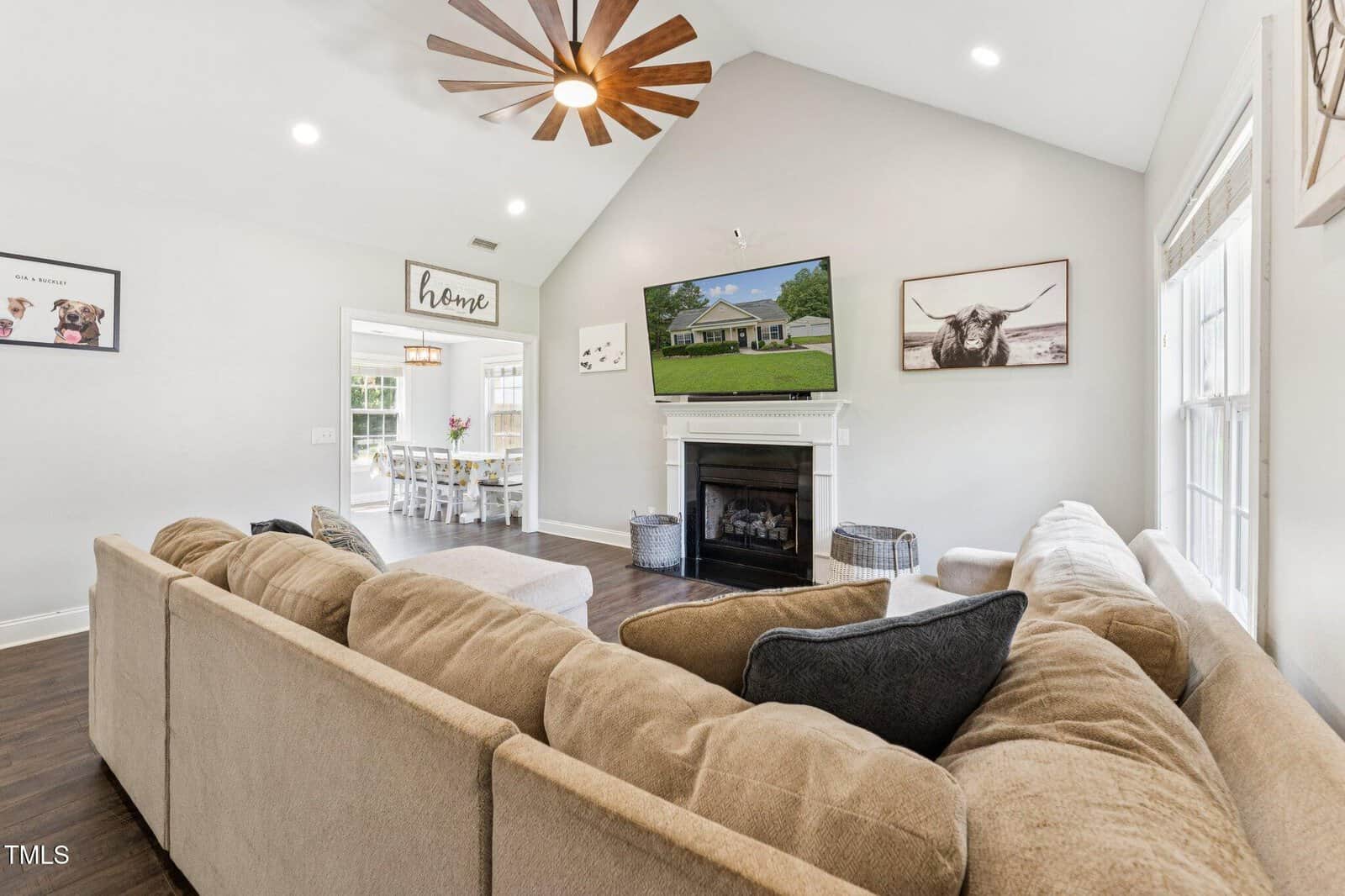 A living room with a beige sectional sofa, a fireplace, a wall-mounted TV, and framed art. Large windows let in natural light; a ceiling fan hangs above. The dining area is visible through a doorway.