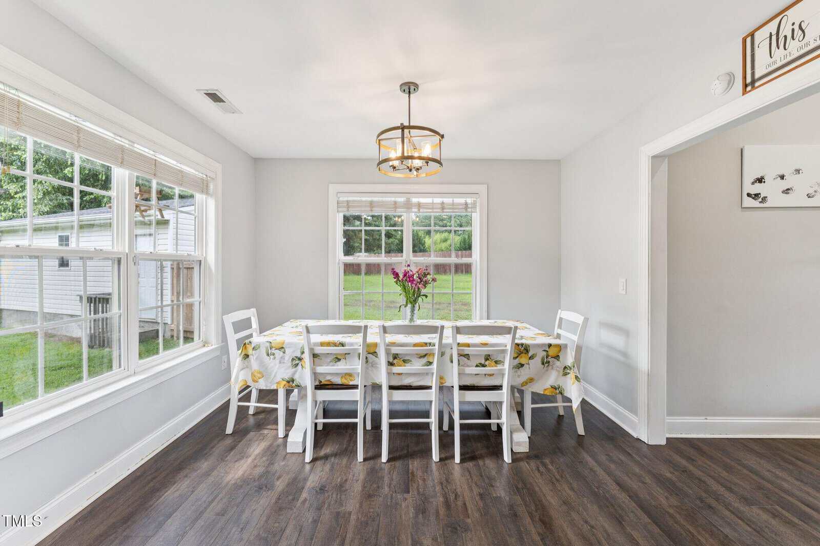 Bright dining room with large windows, white walls, dark wood floor, a long table covered with a floral tablecloth, eight white chairs, and a vase with flowers centered on the table.