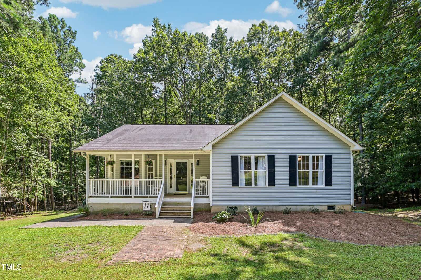 A single-story light gray house at 3642 Wisteria Court features a covered front porch, white railings, black shutters, and three front windows, surrounded by trees and a lawn under the blue Wake Forest sky.